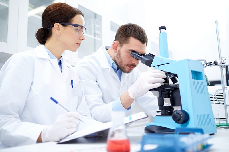 science, chemistry, technology, biology and people concept - young scientists with microscope making test or research in clinical laboratory and taking notes